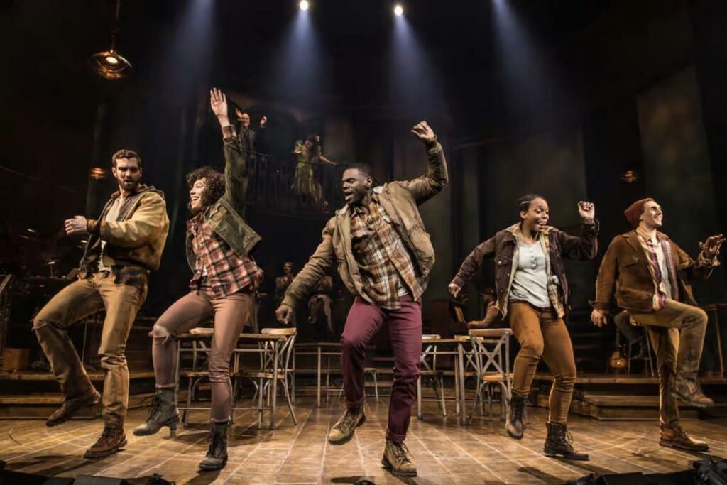 Cena do musical Hadestown na Broadway, mostrando o cenário atmosférico e os músicos no palco durante uma performance deste aclamado musical da Broadway