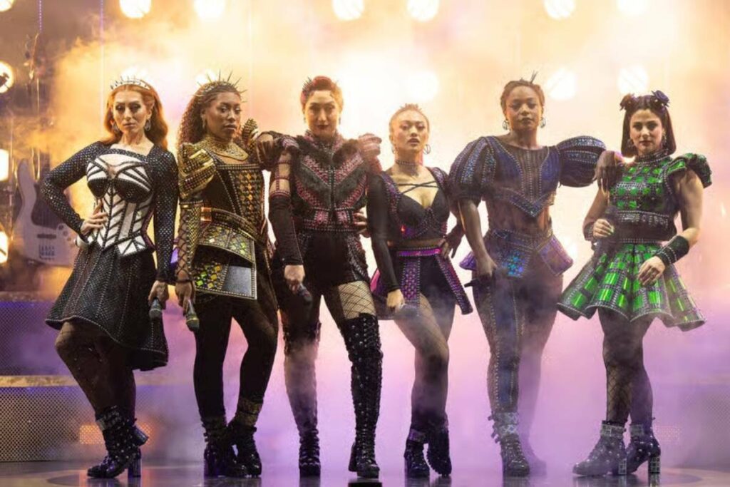 Cena do musical Six na Broadway, mostrando as seis rainhas com seus microfones e figurinos modernos durante uma performance deste inovador musical da Broadway