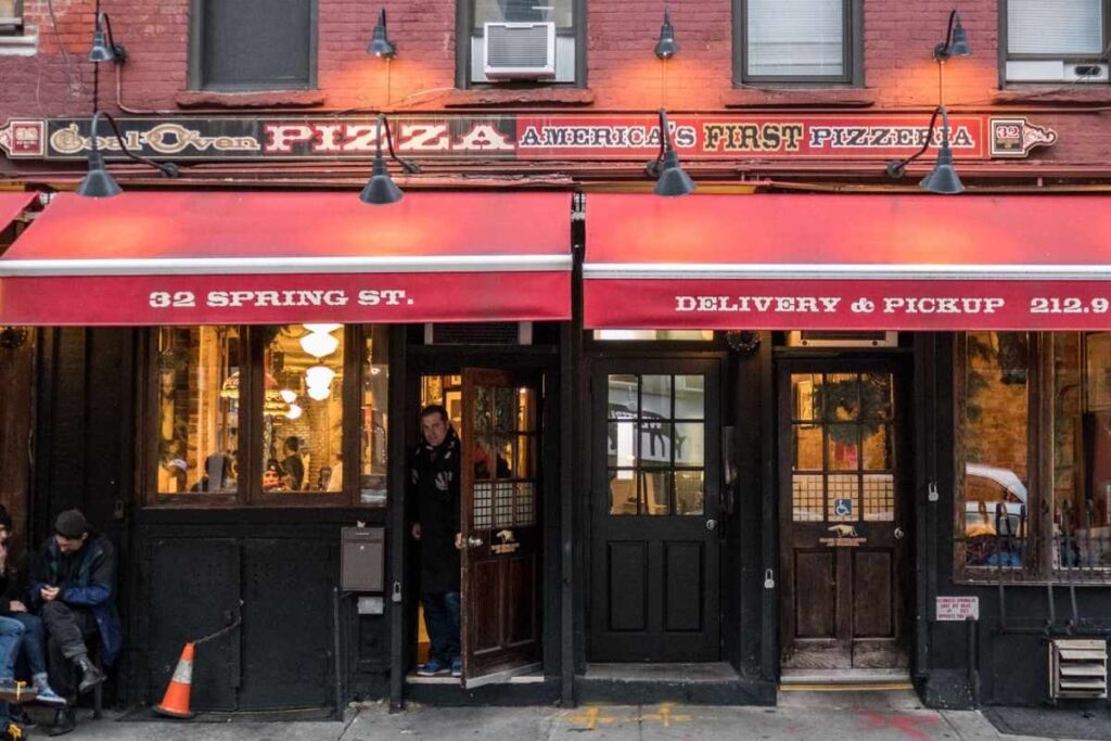 A wood-fired pizza oven, its mouth aglow with a crackling char-kissed crust, sits within a cozy, rustic Italian trattoria. Wafts of yeasty dough and smoked oregano fill the air, mingling with the chatter of families savoring each slice. In the foreground, a freshly baked pie emerges, its surface blistered and glistening, adorned with gooey mozzarella and vibrant tomato sauce. The lighting is warm and intimate, casting a golden glow that enhances the rich, earthy tones of the exposed brick walls and wooden accents. The overall scene evokes the nostalgic charm of a traditional Neapolitan pizzeria, inviting the viewer to step inside and indulge in a truly authentic pizza forno a carvão experience. Lombardi's, a primeira Pizzaria em Nova York