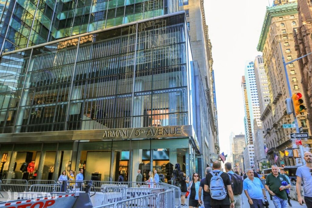 A bustling urban scene on Quinta Avenida, New York City. Towering skyscrapers flank the grand avenue, their glass facades gleaming in the afternoon sunlight. Shoppers stroll along the wide sidewalks, admiring the luxury storefronts and window displays. Elegant townhouses and hotels line the street, with ornate architectural details. A warm, golden light filters through the trees, casting a welcoming glow over the energetic pedestrian traffic. Chic cafes and boutiques beckon, inviting visitors to indulge in the quintessential Quinta Avenida shopping experience. The scene exudes a sense of sophistication, high fashion, and the vibrant pulse of the city. Loja da Armani na Quinta Avenida