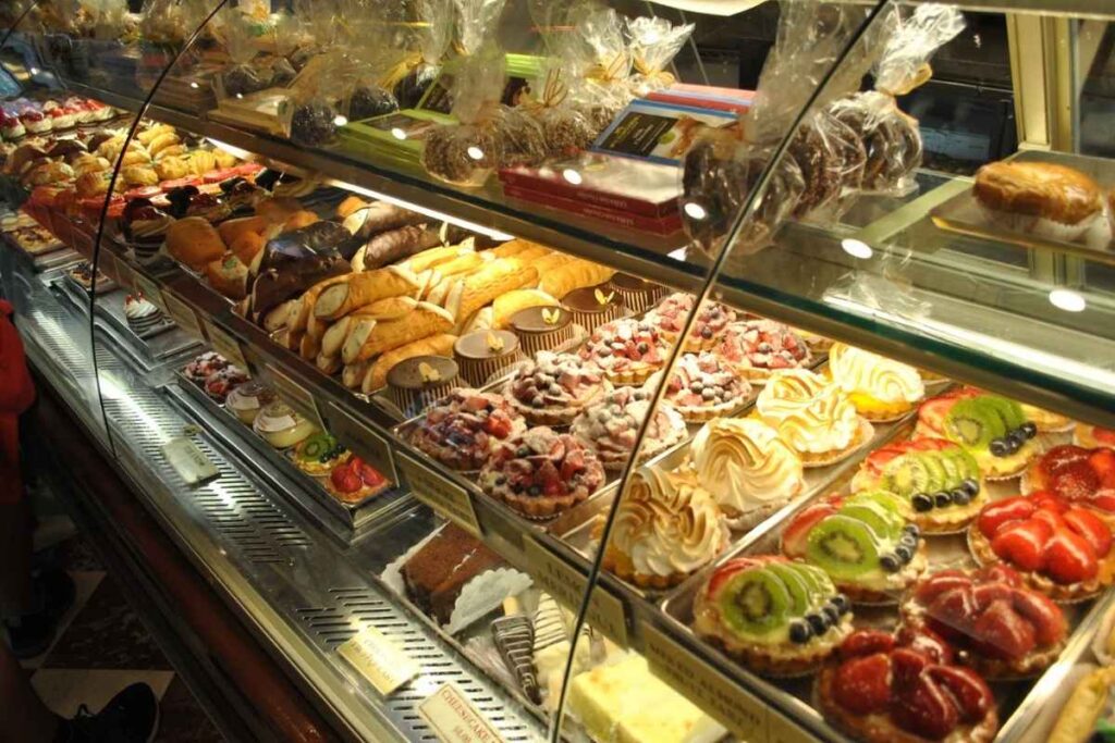 A mouthwatering display of traditional Brazilian doces, including luscious brigadeiros, creamy pudim de leite, and flaky pastel de nata, arranged artfully on a marble countertop. The sweet treats are bathed in soft, warm lighting, casting gentle shadows and highlighting their rich, velvety textures. In the background, a blurred cafe scene, with the aroma of freshly brewed coffee wafting through the air, creating a cozy, inviting atmosphere. The overall composition is elegant and sophisticated, perfectly capturing the essence of an indulgent, quintessential Brazilian dessert experience. Balcão de doces na Ferrara Bakery & Café em Little Italy