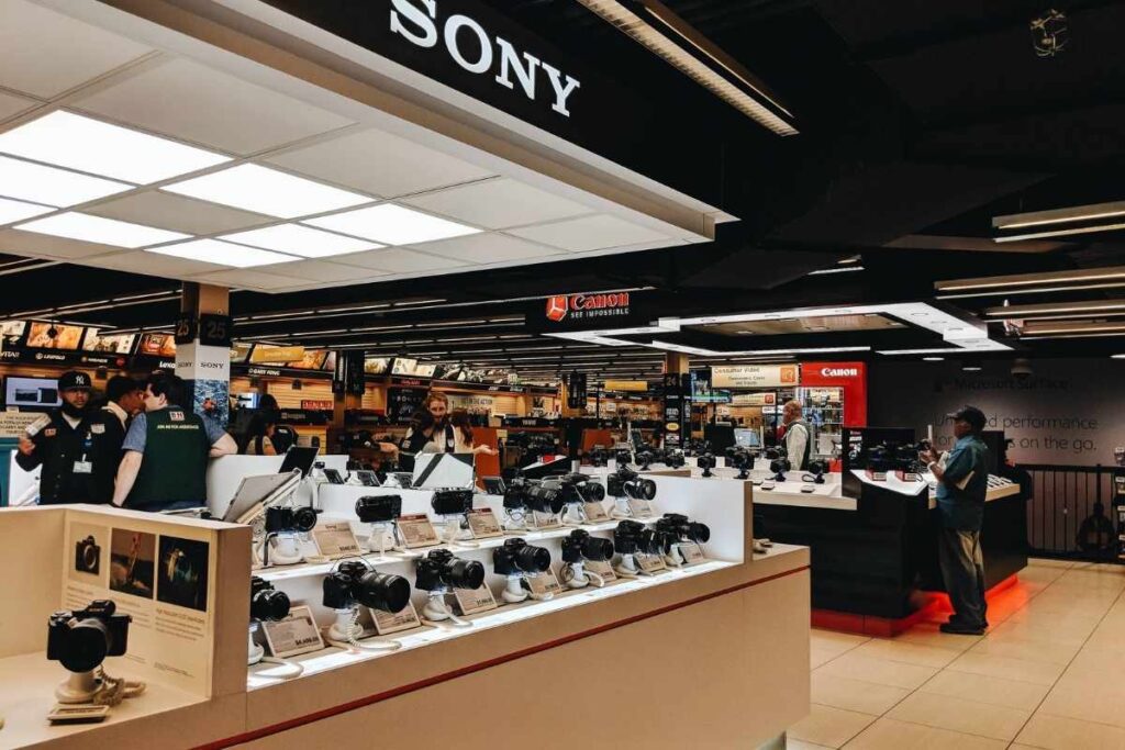 A bustling cityscape in New York City, showcasing a diverse array of modern electronics stores and gadget hubs. In the foreground, sleek display windows feature the latest smartphones, tablets, and wearable technology, illuminated by warm, inviting lighting. The middle ground reveals busy shoppers perusing the aisles, examining the latest tech innovations. In the background, the iconic skyscrapers of Manhattan rise, creating a dynamic, cosmopolitan atmosphere. The scene conveys a sense of excitement and discovery, capturing the thrill of shopping for the latest electronic marvels in one of the world's most vibrant and technologically advanced cities. B&H Photo Video, primeira parada para compra de eletrônicos e fotografia em Nova York