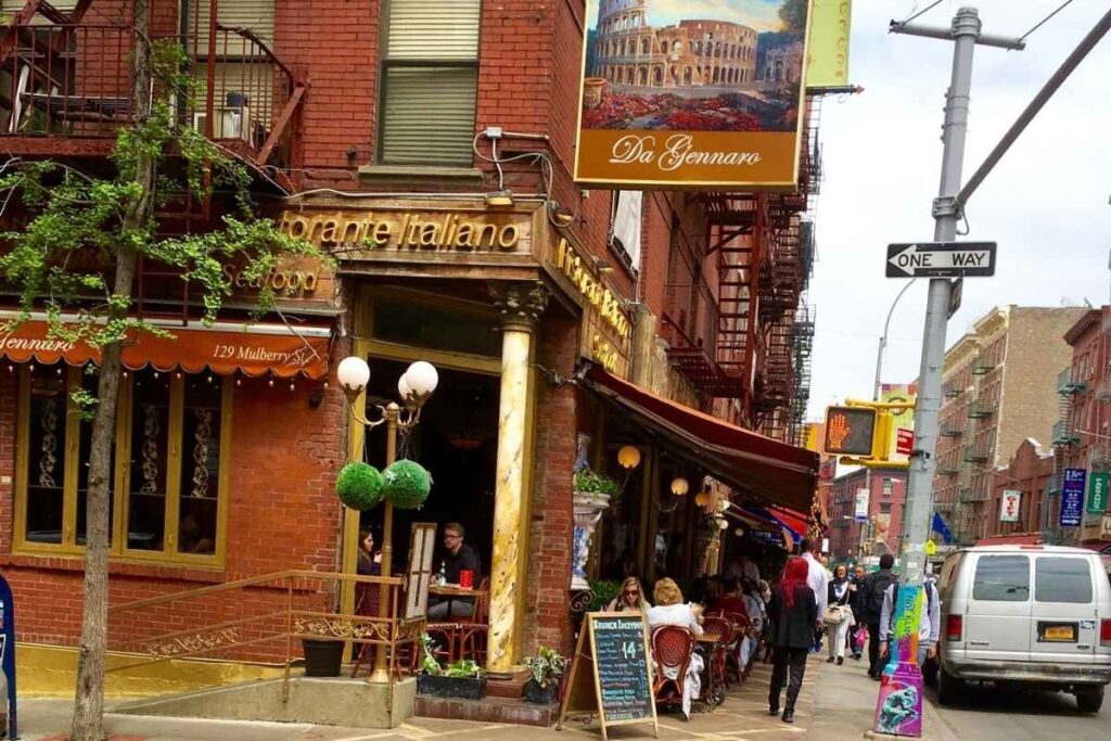 A bustling street in the heart of Little Italy, New York, illuminated by the warm glow of cafe lights. In the foreground, a small group of friends enjoying a lively conversation over a delectable spread of authentic Italian dishes, from steaming plates of pasta to crisp, wood-fired pizzas. In the middle ground, a family-owned trattoria with a charming, old-world ambiance, its windows framed by lush greenery and vibrant floral displays. In the background, the iconic brownstone buildings and cobblestone streets that give this neighborhood its timeless, neighborhood charm. The scene is bathed in a soft, golden light, capturing the inviting, convivial atmosphere that has made Little Italy a renowned culinary destination. Da Gennaro, um dos melhores Restaurantes em Little Italy
