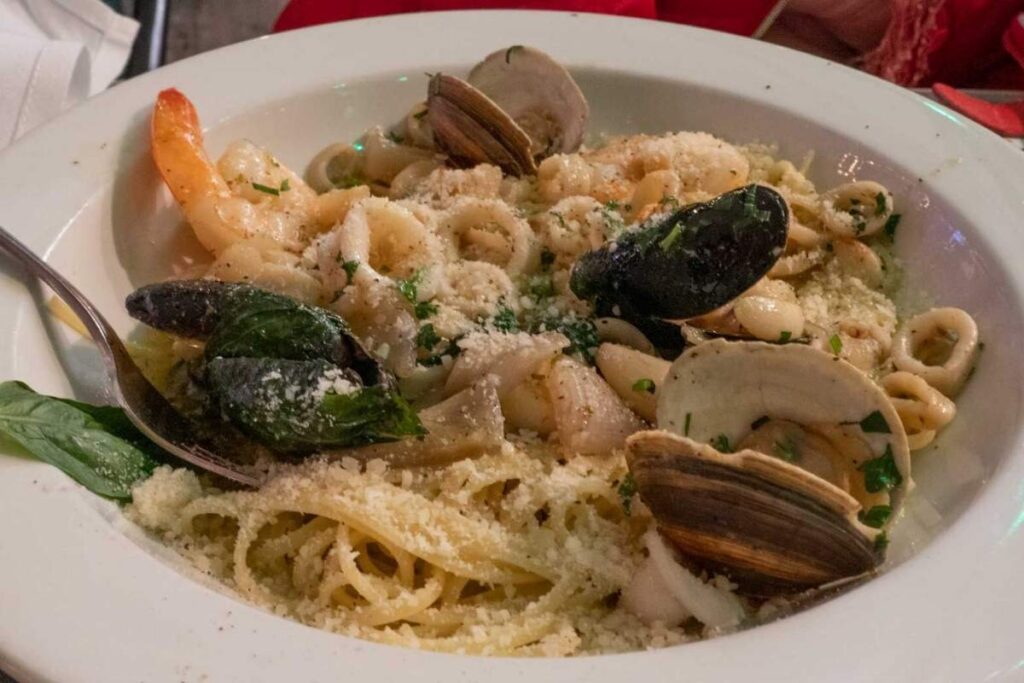 Delectable plate of fresh, hand-crafted pasta in a classic Italian trattoria setting. Foreground showcases a mound of al dente penne in a rich, velvety tomato-based sauce, topped with grated Parmesan and a sprinkle of fragrant basil. Middle ground features a crisp green salad and a glass of robust red wine. Background depicts a cozy, warm-lit dining room with exposed brick walls, wooden beams, and checkered tablecloths, evoking the inviting ambiance of a beloved neighborhood eatery in the heart of Little Italy. Massa com frutos do mar no IL Cortile