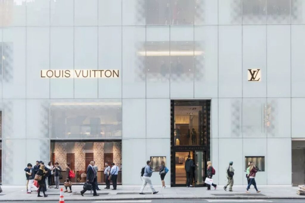 A luxurious five-story Louis Vuitton flagship store stands prominently on the iconic Fifth Avenue in New York City. The building's distinctive architecture features sleek glass facades, elegant stone detailing, and the brand's signature monogram pattern. The store's entrance is flanked by tall palm trees, and the sidewalk is bustling with well-heeled shoppers. Bright, even lighting illuminates the scene, capturing the glamour and sophistication of high-end retail. The image conveys the allure of luxury shopping on one of the world's most famous shopping destinations. Lojas consagradas como Louis Vuitton e Yves Saint Laurent estão na Quinta Avenida