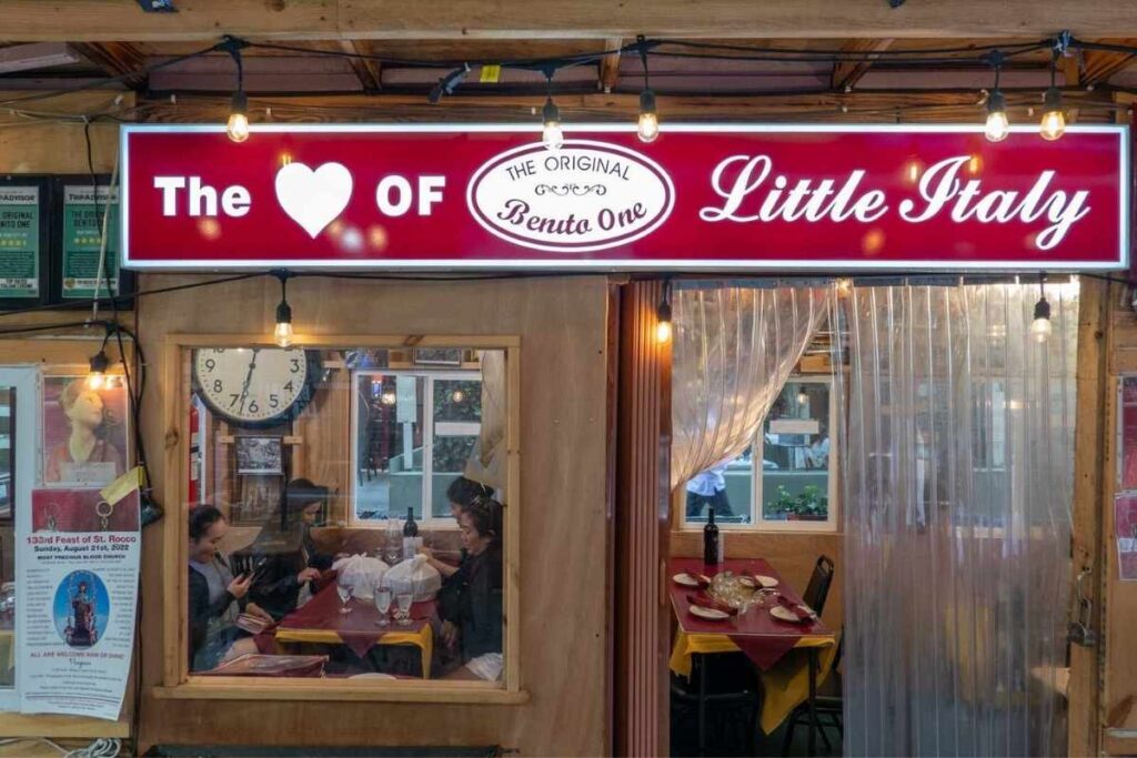 A cozy Italian wine cellar, dimly lit by soft golden lamps, showcasing an assortment of premium vintages. In the foreground, an elegant display of aperitifs and local gourmet delicacies, arranged on a rustic wooden table, inviting the viewer to indulge in a complete sensory experience. The middle ground features towering shelves, lined with carefully curated bottles, each with a unique label and story to tell. In the background, a warm, textured brick wall creates a sense of timeless tradition, while large windows allow natural light to filter in, casting a gentle glow over the scene. The overall atmosphere evokes a feeling of old-world charm and sophisticated indulgence, perfectly capturing the essence of a Little Italy dining experience.