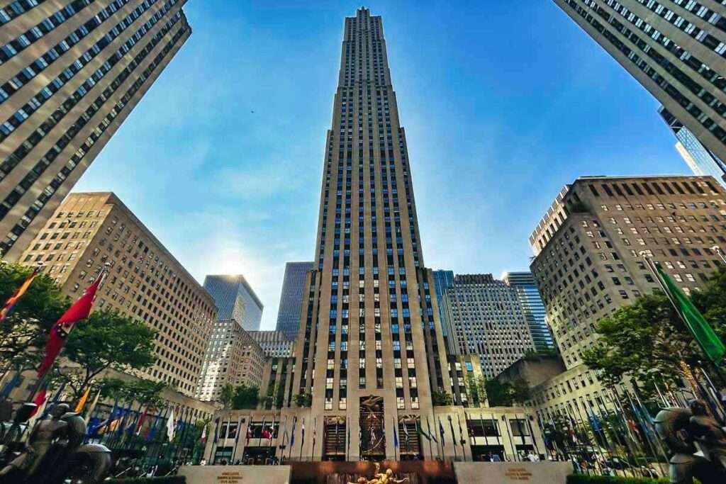 A stunning Art Deco skyline in New York City, with the iconic Chrysler Building standing tall against a clear blue sky. In the foreground, ornate stone facades and geometric designs adorn the facades of nearby skyscrapers, casting long shadows across the bustling streets below. The mid-ground features the sweeping curves and gleaming chrome of the Rockefeller Center, its observation decks offering breathtaking views of the city. Soft, diffused lighting creates a warm, elegant atmosphere, emphasizing the architectural details and the graceful lines of the buildings. The overall composition conveys a sense of grandeur, sophistication, and the Golden Age of New York City. Rockefeller Center