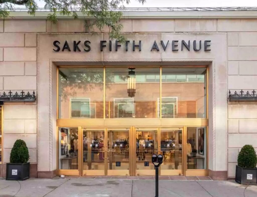 A grand, neoclassical façade with intricate columns, ornate entryways, and gleaming glass displays. Elegant shoppers stroll along the bustling sidewalk, past lush potted plants and flags waving in a gentle breeze. Warm, directional lighting casts a soft glow, highlighting the building's regal architecture and the refined luxury of the Saks Fifth Avenue storefront. In the distance, the towering skyscrapers of New York City's iconic skyline provide a dramatic backdrop, conveying the prestige and glamour of this renowned department store on the famous Fifth Avenue. A Saks Fifth Avenue