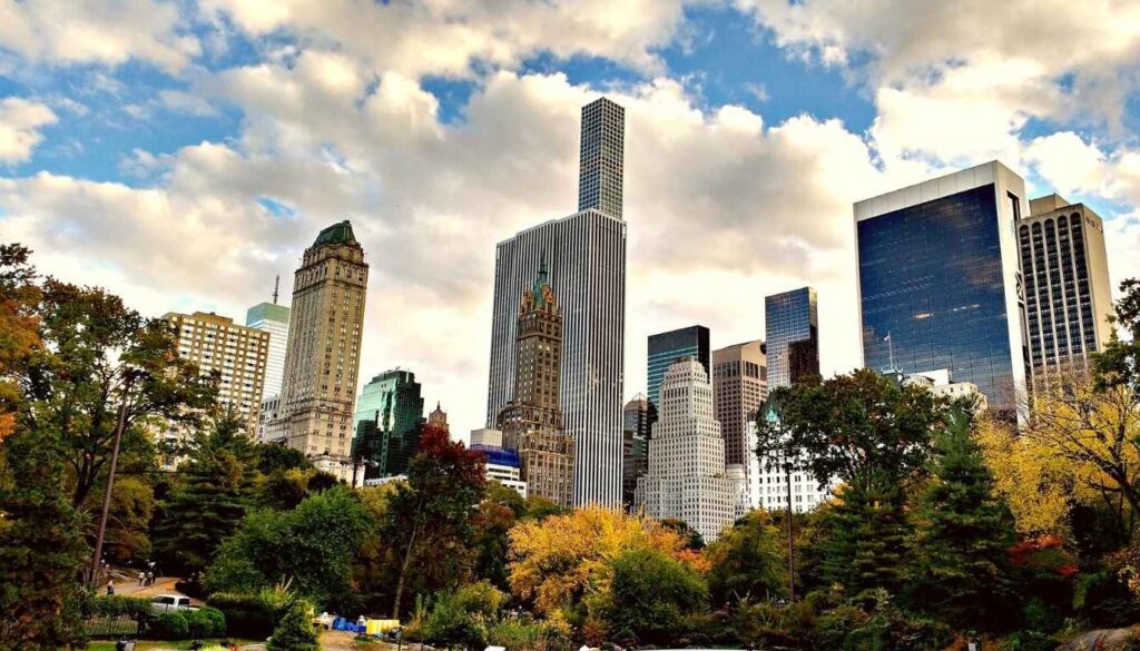 A serene and expansive vista of Central Park, captured on a mild autumn day. In the foreground, a winding path flanked by lush greenery and vibrant fall foliage invites exploration. The middle ground features the iconic Bethesda Fountain, its cascading waters surrounded by ornate architecture and stately trees. In the distance, the towering Manhattan skyline stands in contrast, a testament to the park's role as an urban oasis. Soft, diffused lighting filters through the clouds, casting a warm glow over the scene and accentuating the park's natural beauty. The overall composition conveys a sense of tranquility and wonder, perfectly capturing the essence of a stroll through this beloved New York landmark. Vista do Skyline de Manhattan a partir do Central Park