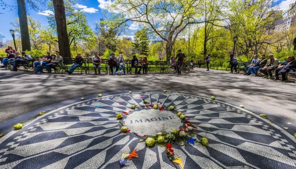 A lush, vibrant field of ripe strawberries stretching across the frame, their ruby-red hues glistening in the warm sunlight. In the distance, a mosaic of John Lennon's iconic face, "Imagine" etched into the ground, creating a serene and contemplative atmosphere. Surrounding the mosaic, verdant trees sway gently, their leaves casting a dappled pattern of light and shadow over the scene. The composition is framed by a warm, golden hour glow, the image captured through a wide-angle lens to showcase the expansive, dreamlike quality of this peaceful, idyllic setting. Strawberry Fields é o cantinho dedicado a John Lennon no Central Park