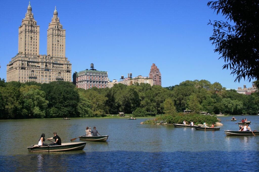 A serene and picturesque lake in Central Park, with the iconic Loeb Boathouse nestled along the shoreline. Softly rippling waters reflect the lush foliage and towering trees that line the banks. Elegant wooden rowboats and traditional Venetian-style gondolas gently drift across the tranquil surface, their passengers taking in the peaceful ambiance. Warm, golden sunlight filters through the branches, casting a gentle glow over the scene. The boathouse's classic architecture stands as a charming focal point, its warm brick and weathered wood evoking a timeless elegance. An atmosphere of leisure and relaxation pervades the entire tableau, inviting the viewer to imagine themselves drifting along the lake, immersed in the serene beauty of this iconic New York City landscape. The Lake