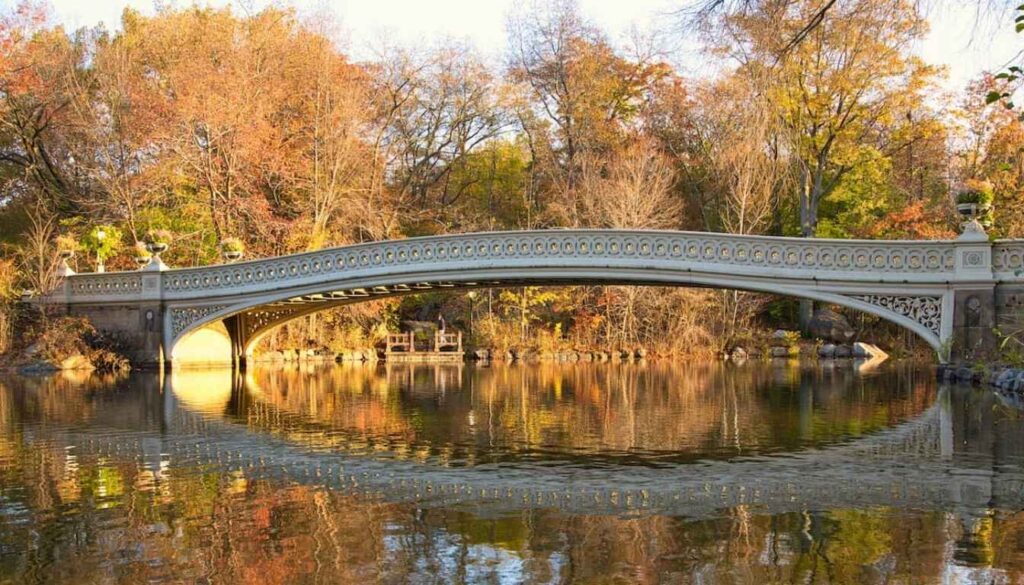 A breathtaking view of the iconic Bow Bridge in New York's Central Park, captured in the warm glow of the golden hour. The graceful stone arch reflects in the calm waters, creating a picturesque scene. In the foreground, a couple strolls hand-in-hand, their silhouettes framed by the elegant curves of the bridge. The middle ground features lush greenery and blossoming trees, adding depth and natural beauty to the composition. In the background, the iconic Manhattan skyline rises, creating a stunning contrast against the serene park setting. The image is rendered with a cinematic, high-resolution quality, inviting the viewer to feel the tranquility and romance of this renowned photographic location. A Bow Bridge é um a das Top 10 Coisas pra Fazer no Central Park