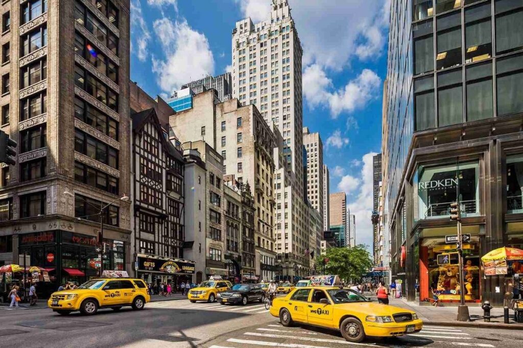 A vibrant and luxurious scene on Fifth Avenue in New York City, showcasing its iconic architecture, high-end shopping, and bustling atmosphere. The foreground features the grand, neo-Renaissance style of the Fifth Avenue buildings, with intricate facades, ornate windows, and towering spires. In the middle ground, people stroll along the sidewalks, window-shopping at the renowned luxury brand stores. The background is dominated by the towering skyscrapers of Midtown Manhattan, creating a dynamic contrast between old and new. Warm, golden lighting illuminates the scene, casting a dreamy, sophisticated ambiance. The image conveys the essence of Fifth Avenue as a hub of luxury, culture, and commerce, inviting the viewer to immerse themselves in the vibrant energy of New York City. Visão geral da Quinta Avenida