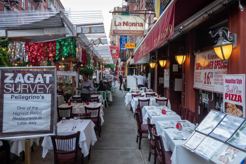 A bustling street in the heart of Little Italy, lined with cozy ristorantes and cafes. In the foreground, a group of diners enjoying a lively alfresco meal, surrounded by the warm glow of string lights and colorful awnings. The middle ground features a mix of classic Italian architecture, with ornate balconies and terracotta roofs. In the background, a towering cathedral stands as a testament to the neighborhood's rich cultural heritage. The scene is bathed in a soft, golden light, creating an inviting and authentic atmosphere that captures the essence of the best restaurants in Little Italy. Restaurante em Little Italy com mesas na calçada