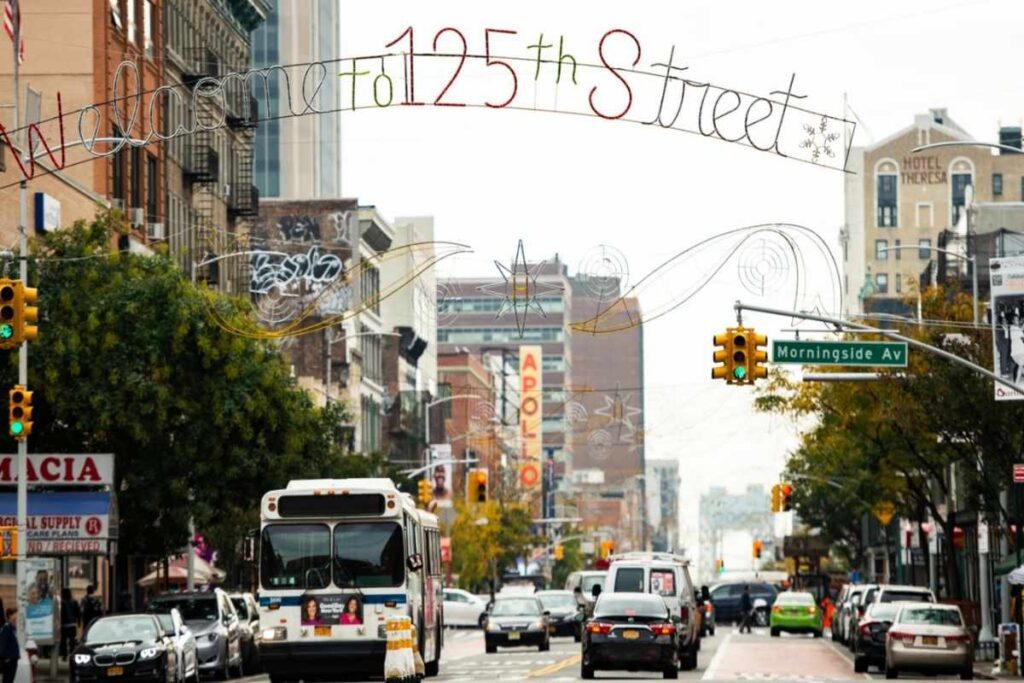 Cena de rua movimentada em Harlem, Nova York, com pessoas caminhando e prédios históricos ao fundo.