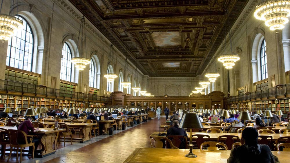Pessoas diversas estudando e lendo dentro de uma grande biblioteca pública de Nova York, com estantes de livros, janelas grandes