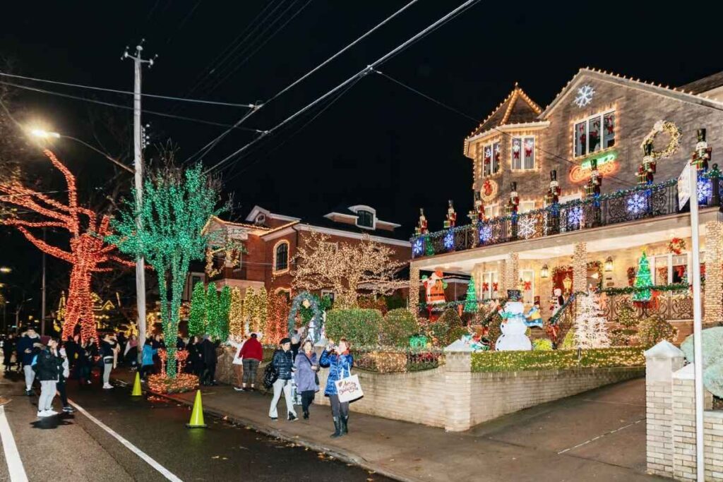 Rua residencial em Dyker Heights, Brooklyn, com casas decoradas para o Natal, pessoas caminhando e árvores iluminadas.
