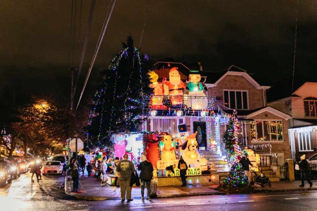 Rua residencial em Dyker Heights decorada com luzes de Natal coloridas e pessoas caminhando e admirando as decorações natalinas.