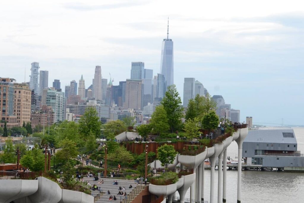 Vista aérea do parque flutuante Little Island em Nova York, mostrando áreas verdes, caminhos e pessoas, com o rio Hudson e o horizonte da cidade ao fundo.